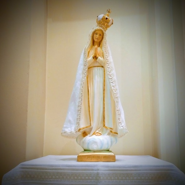 Statue of the Blessed Virgin Mary in white clothing embroidered with gold details. Her hands are pressed together in prayer and her head tilts slightly downward and to the right.