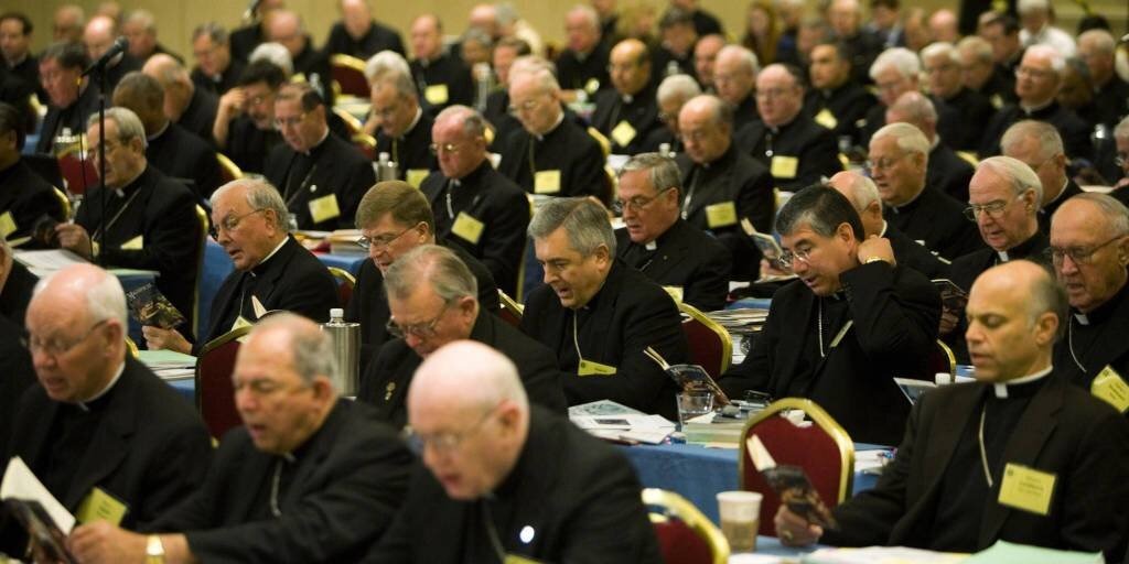Rows of Catholic bishops in black clerical attire seated at conference tables, reading documents and booklets during a large assembly.
